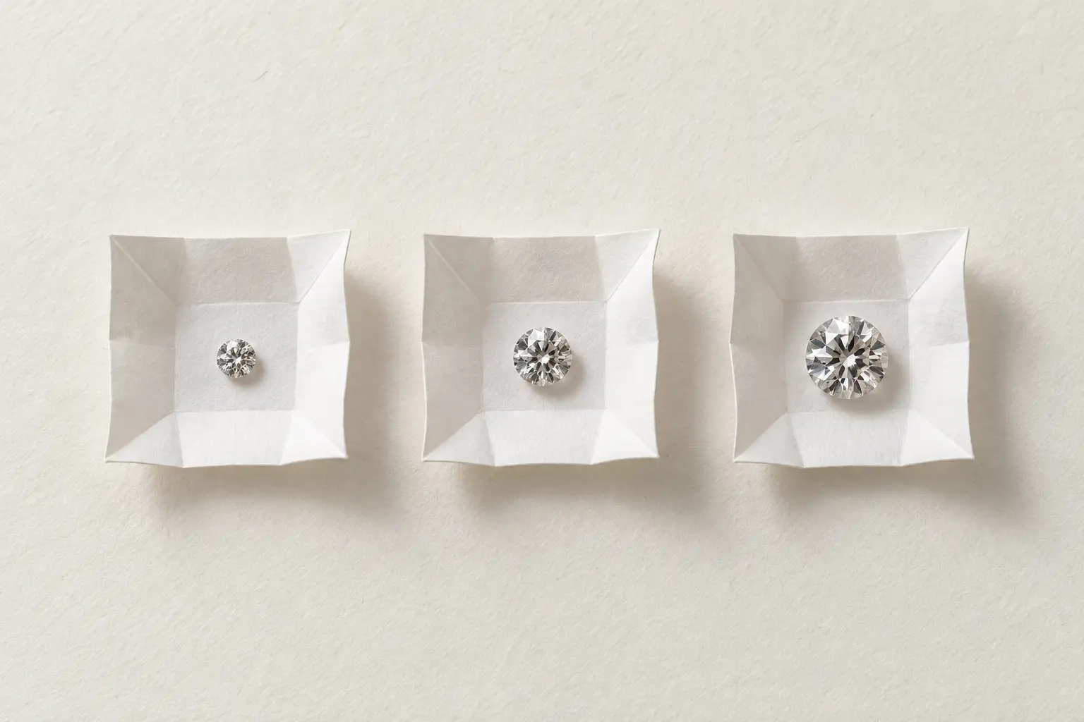 Three loose round diamonds of increasing size sit in a row on three small folds of white paper.