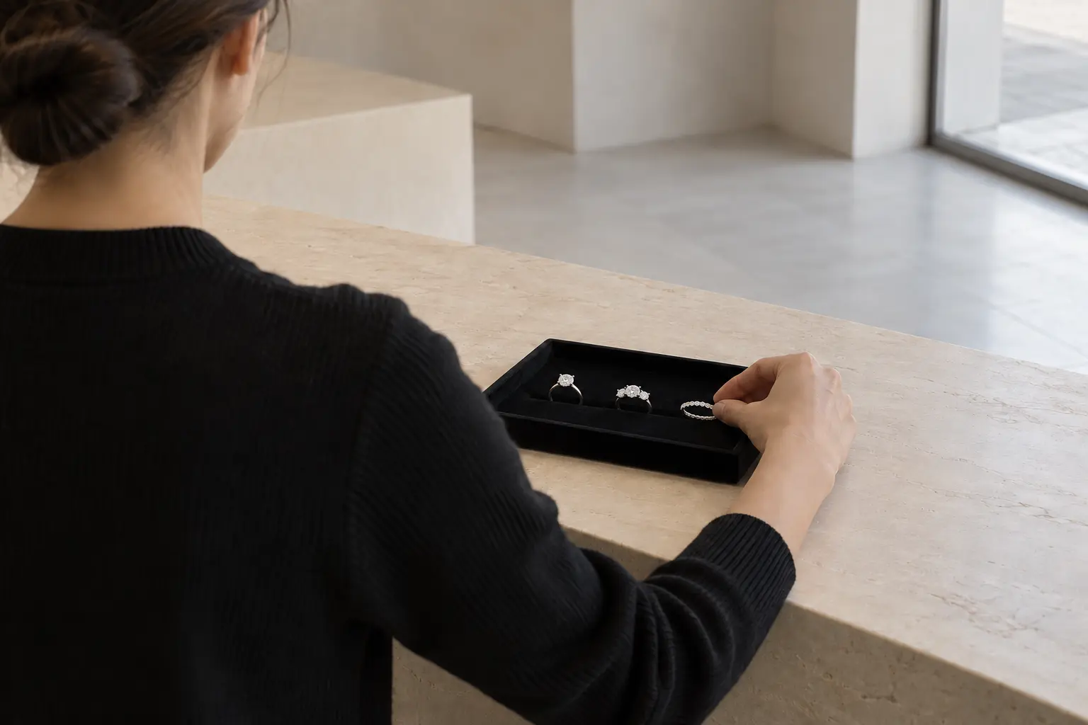 A jeweler, seen from behind, places the third of three different rings in a black velvet tray on a pale stone counter in a quiet boutique.